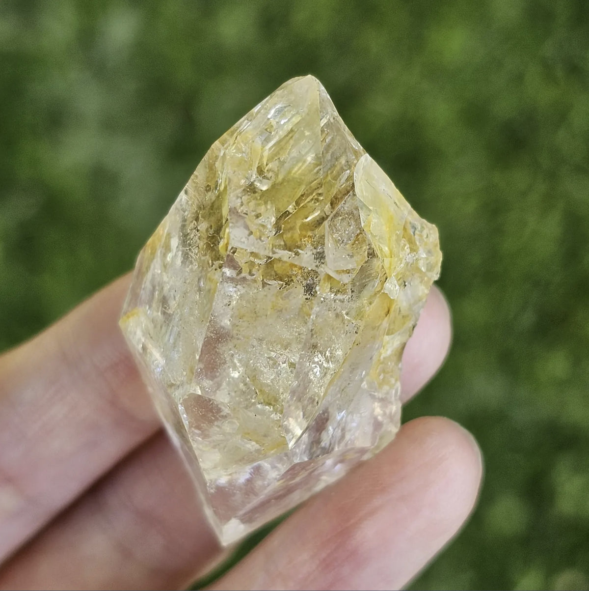 A hand holding a clear, double-terminated New York Herkimer Diamond quartz crystal with natural geometric facets and an internal rainbow reflection against a soft green grass background.