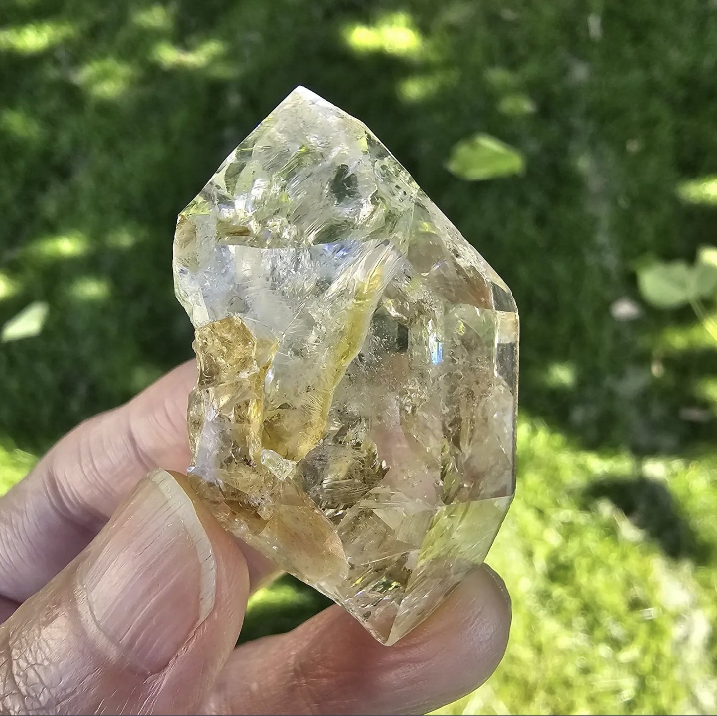 A hand holding a clear, double-terminated New York Herkimer Diamond quartz crystal with natural geometric facets and an internal rainbow reflection against a soft green grass background.