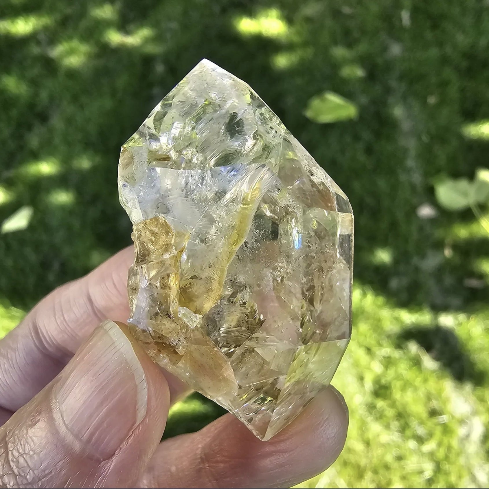 A hand holding a clear, double-terminated New York Herkimer Diamond quartz crystal with natural geometric facets and an internal rainbow reflection against a soft green grass background.