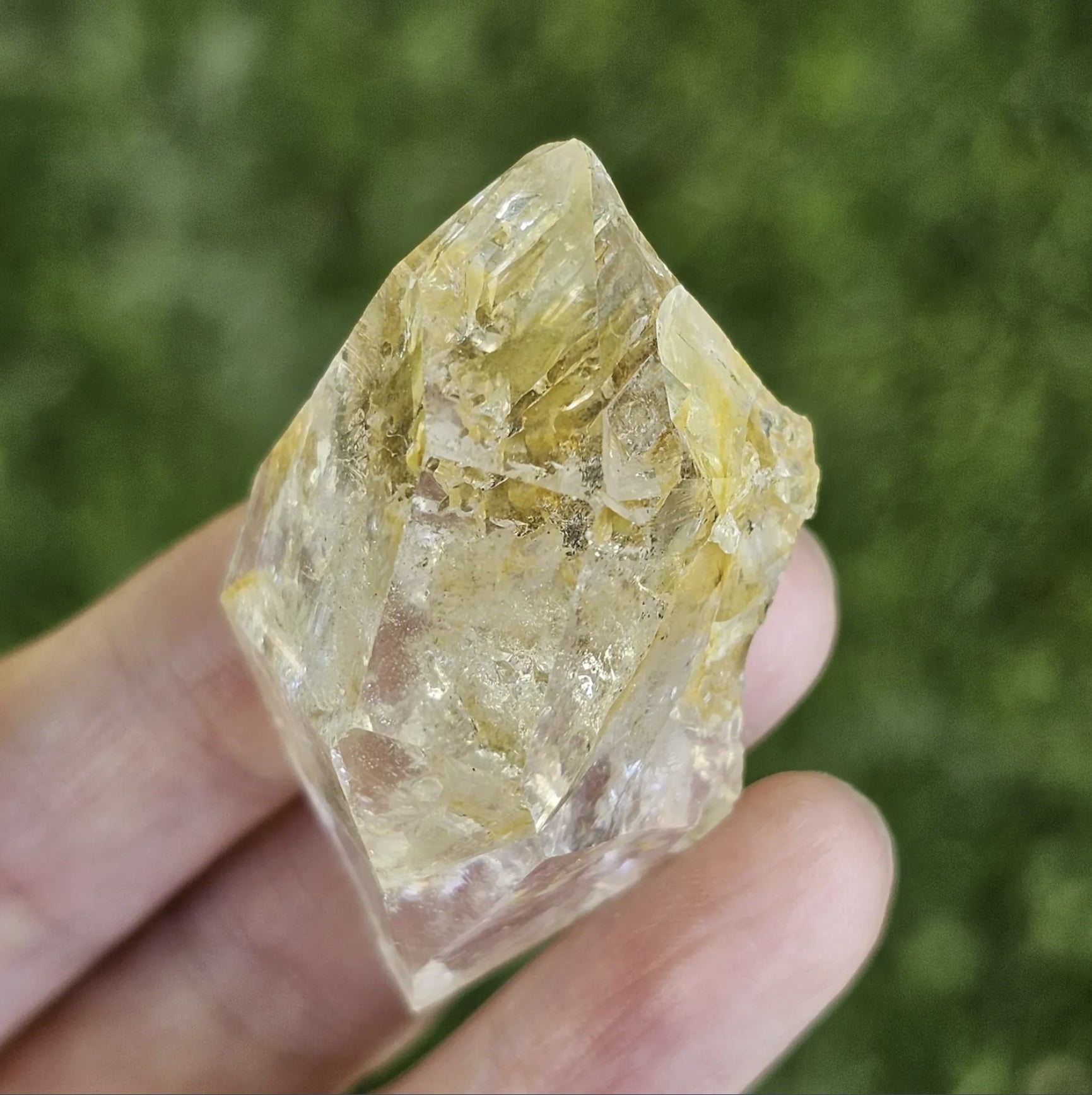 A hand holding a clear, double-terminated New York Herkimer Diamond quartz crystal with natural geometric facets and an internal rainbow reflection against a soft green grass background.