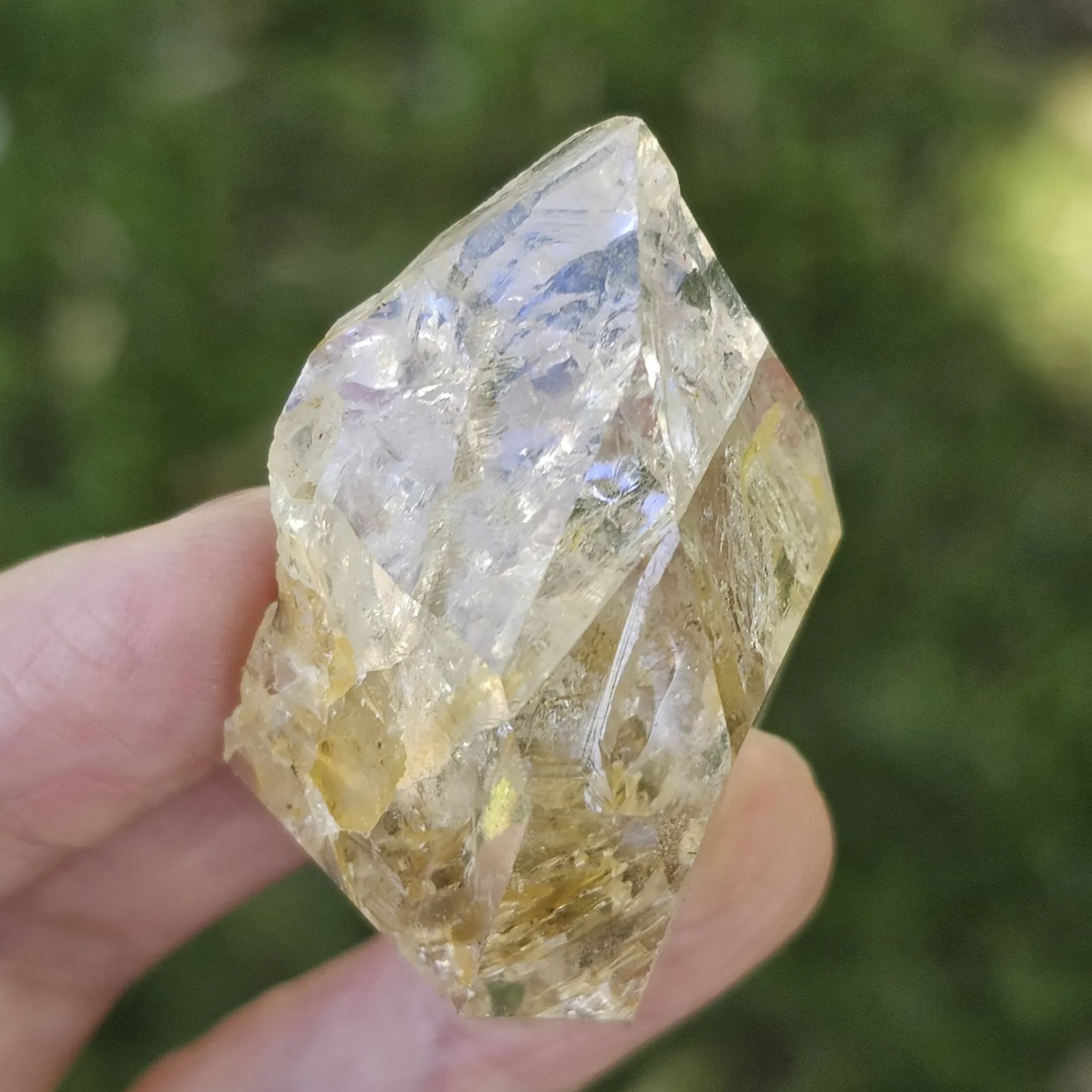 A hand holding a clear, double-terminated New York Herkimer Diamond quartz crystal with natural geometric facets and an internal rainbow reflection against a soft green grass background.