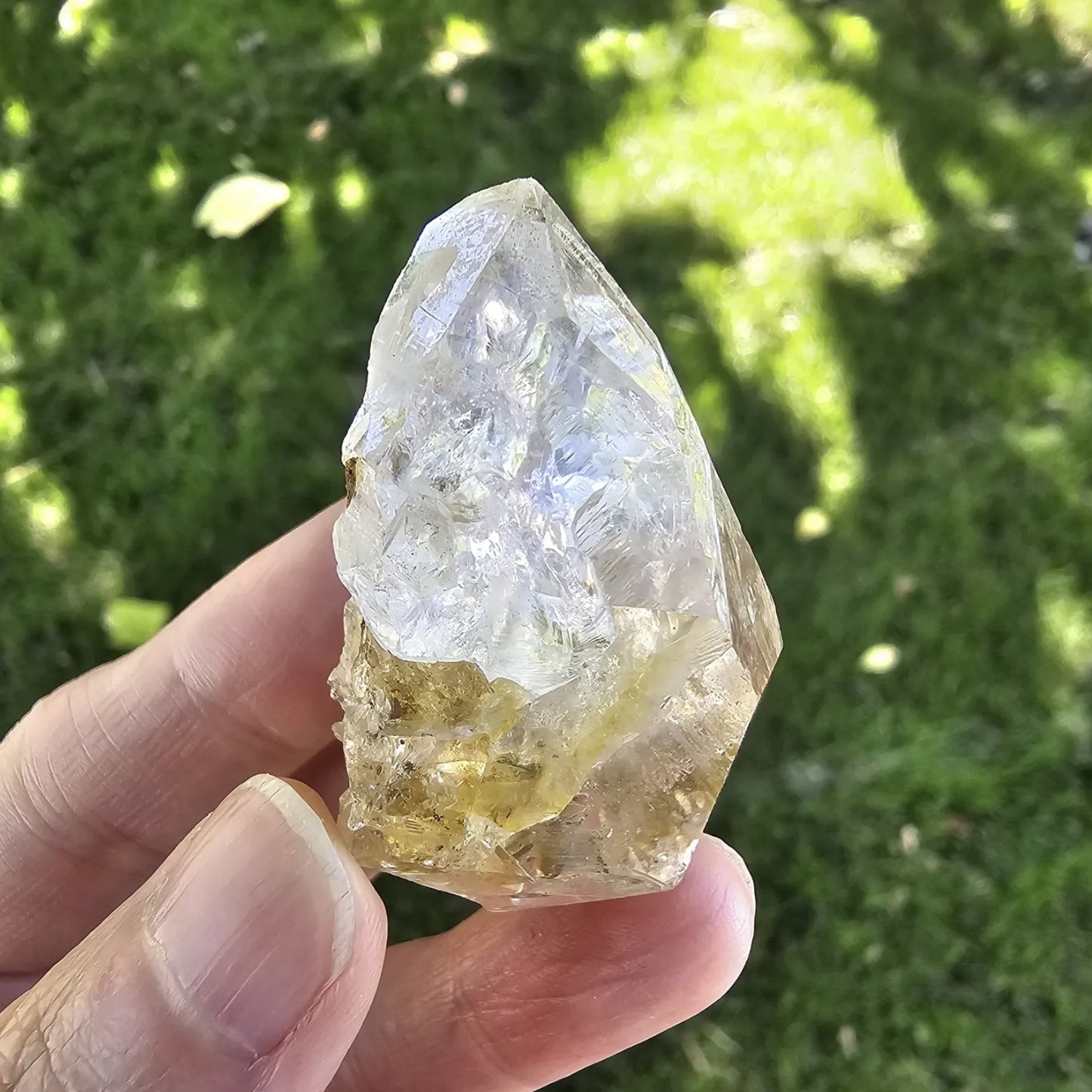 A hand holding a clear, double-terminated New York Herkimer Diamond quartz crystal with natural geometric facets and an internal rainbow reflection against a soft green grass background.