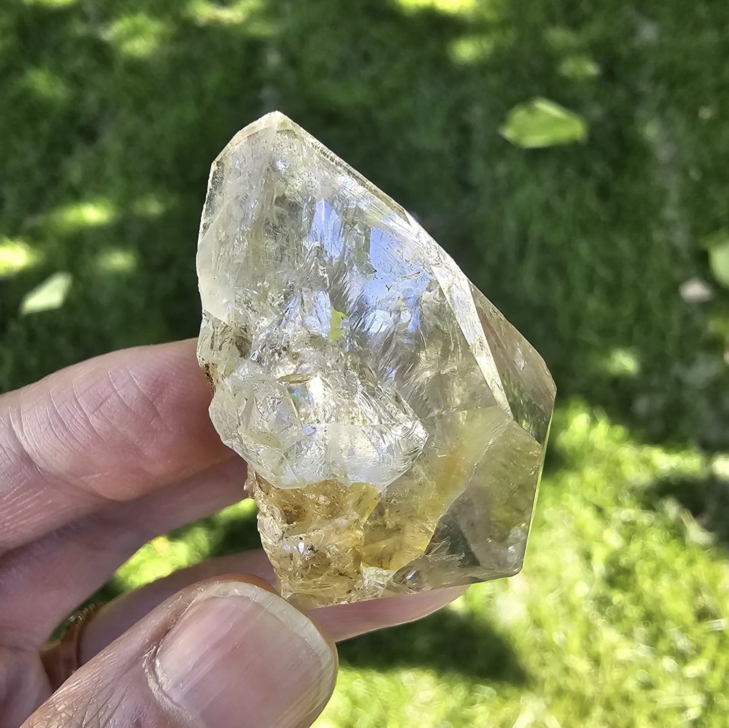 A hand holding a clear, double-terminated New York Herkimer Diamond quartz crystal with natural geometric facets and an internal rainbow reflection against a soft green grass background.