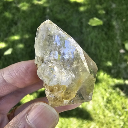 A hand holding a clear, double-terminated New York Herkimer Diamond quartz crystal with natural geometric facets and an internal rainbow reflection against a soft green grass background.