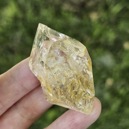 A hand holding a clear, double-terminated New York Herkimer Diamond quartz crystal with natural geometric facets and an internal rainbow reflection against a soft green grass background.