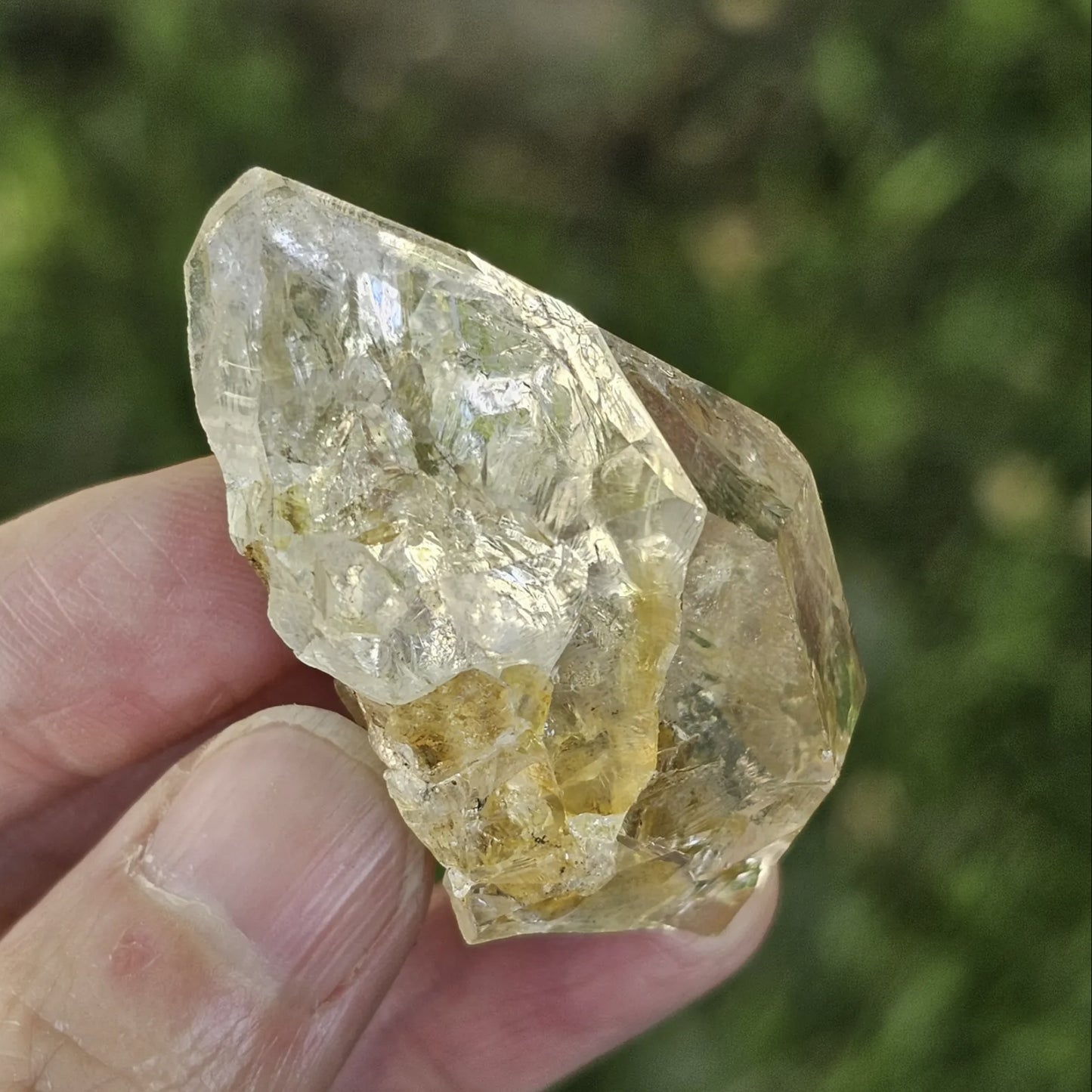 A hand holding a clear, double-terminated New York Herkimer Diamond quartz crystal with natural geometric facets and an internal rainbow reflection against a soft green grass background.