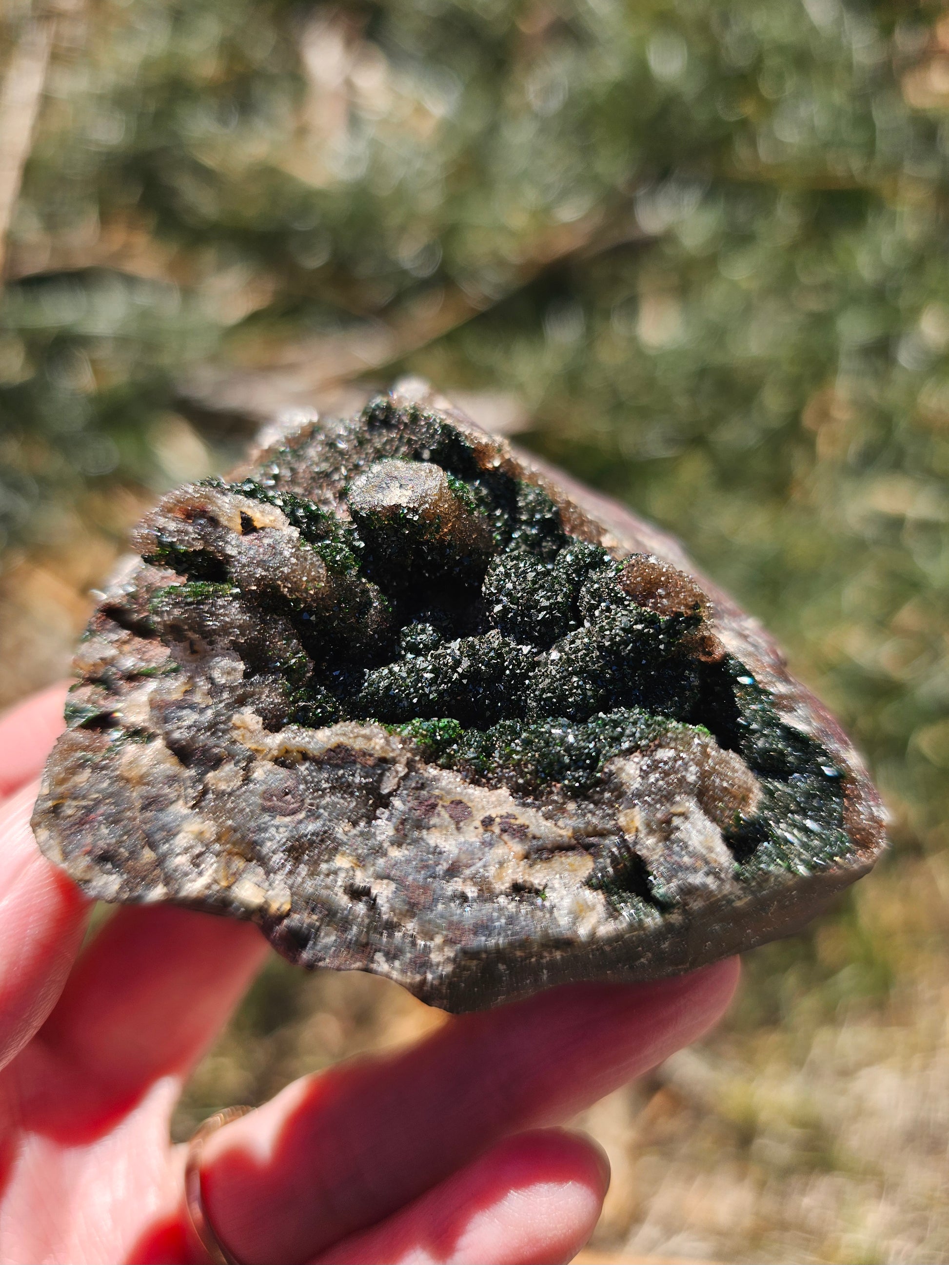 A hand holding a 109g Congo crystal specimen to show its 2.5 inch size, displaying the weight and substantial grounding presence of the Hematite and Duftite minerals.