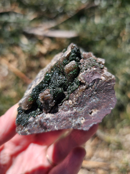 A hand holding a 109g Congo crystal specimen to show its 2.5 inch size, displaying the weight and substantial grounding presence of the Hematite and Duftite minerals.