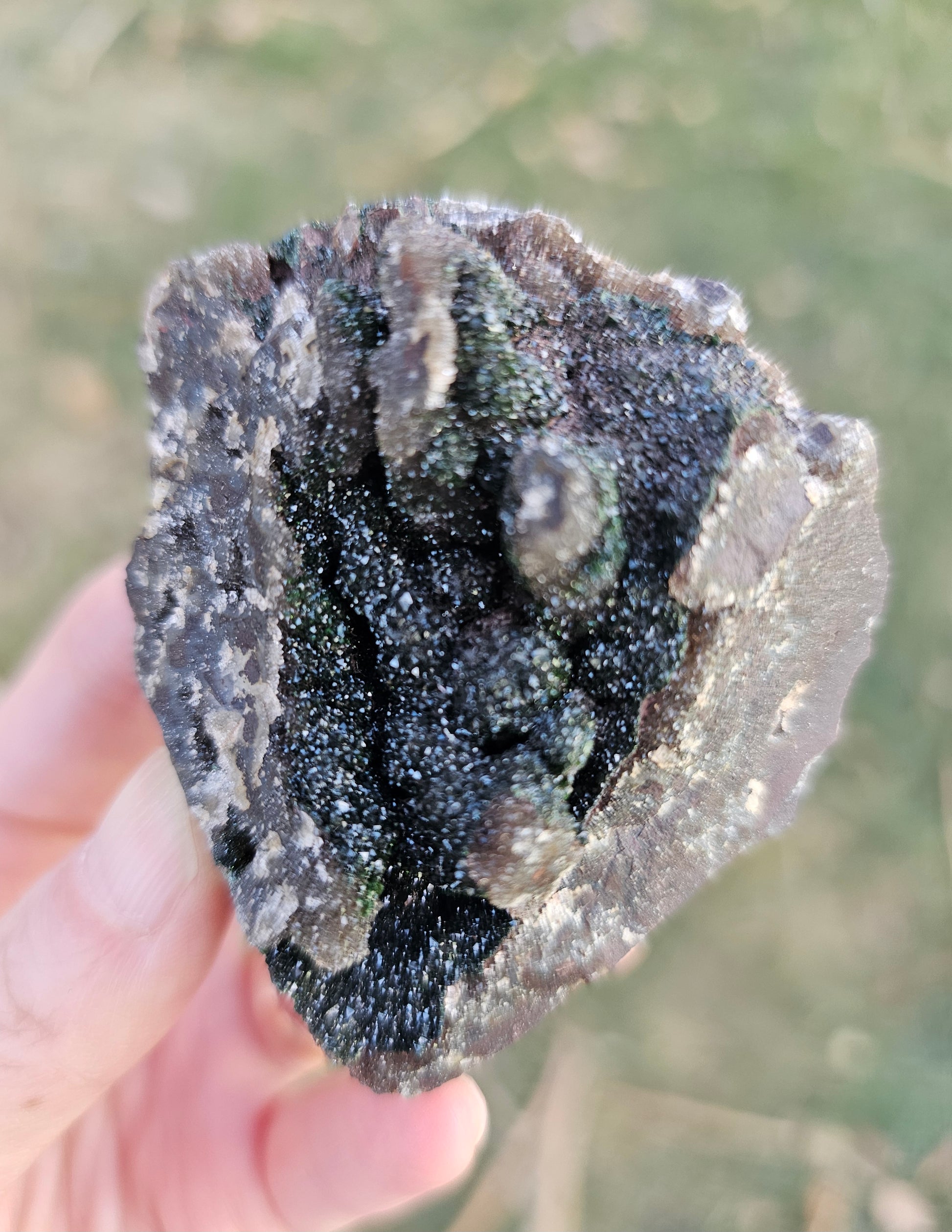 A hand holding a 109g Congo crystal specimen to show its 2.5 inch size, displaying the weight and substantial grounding presence of the Hematite and Duftite minerals.