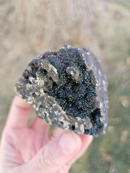 A hand holding a 109g Congo crystal specimen to show its 2.5 inch size, displaying the weight and substantial grounding presence of the Hematite and Duftite minerals.