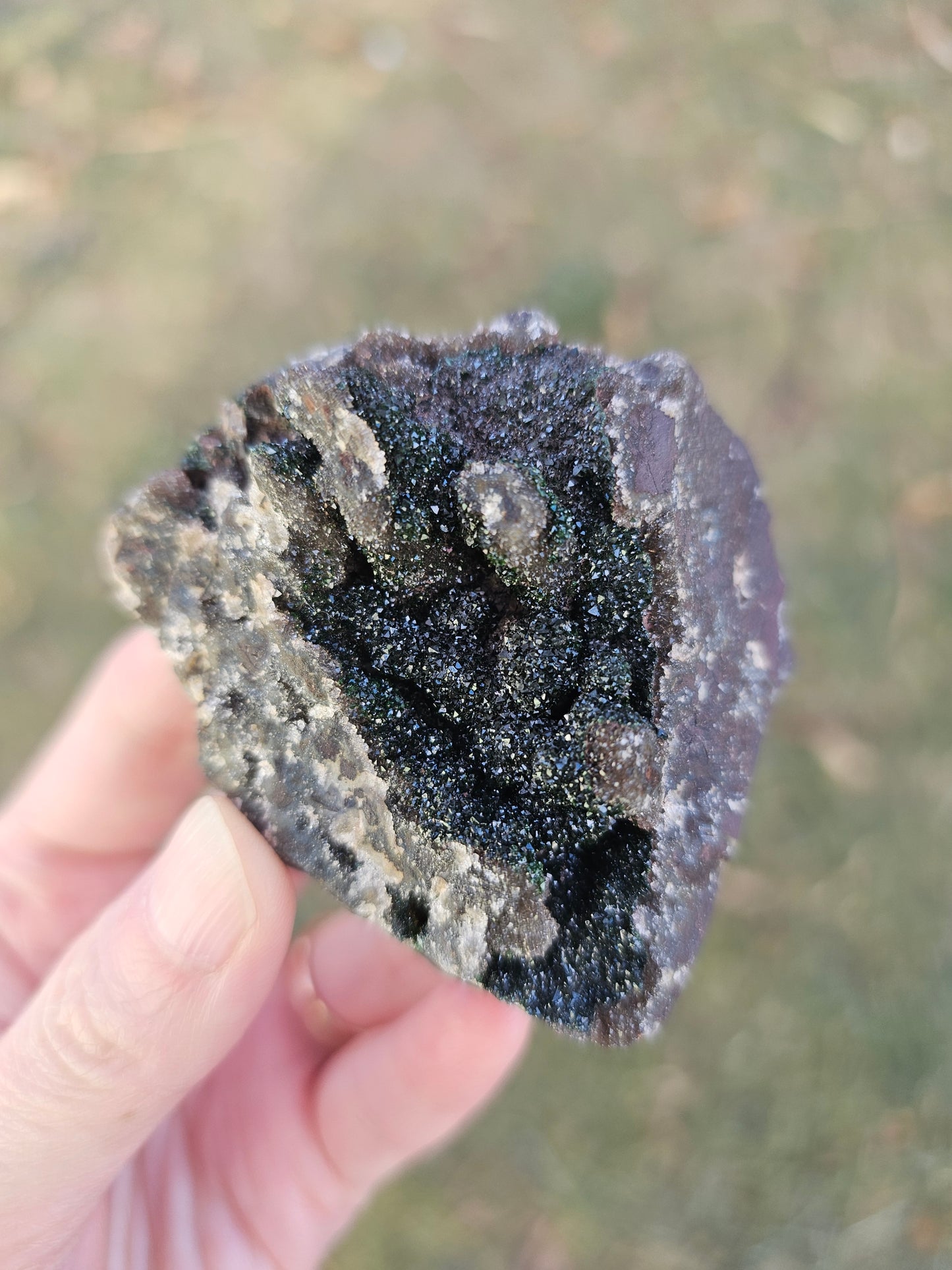 A hand holding a 109g Congo crystal specimen to show its 2.5 inch size, displaying the weight and substantial grounding presence of the Hematite and Duftite minerals.