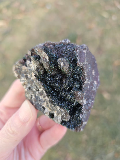 A hand holding a 109g Congo crystal specimen to show its 2.5 inch size, displaying the weight and substantial grounding presence of the Hematite and Duftite minerals.