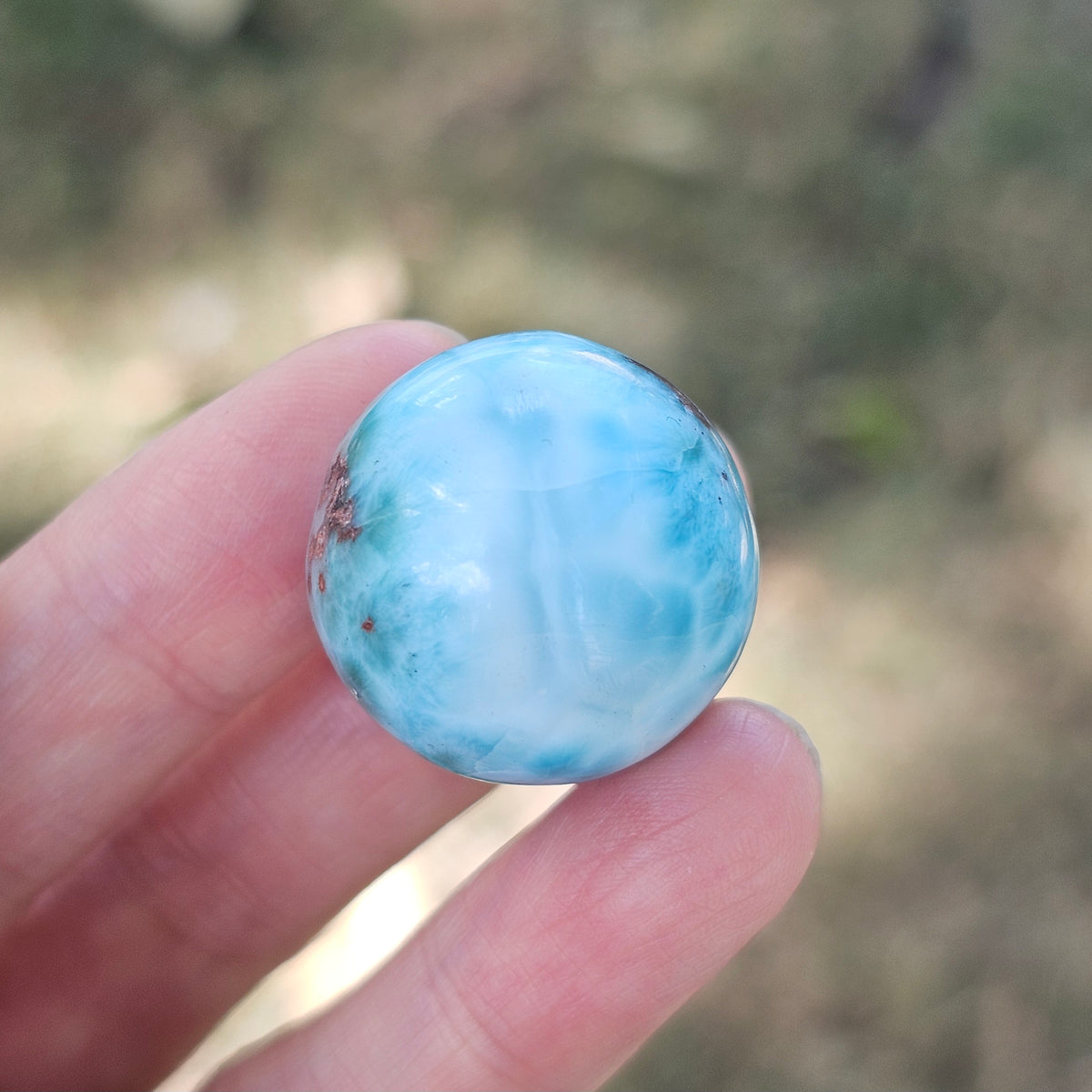 Grade AA natural blue Larimar sphere 16.8g 0.75 inch held in hand showing ocean wave patterns and natural hematite spots