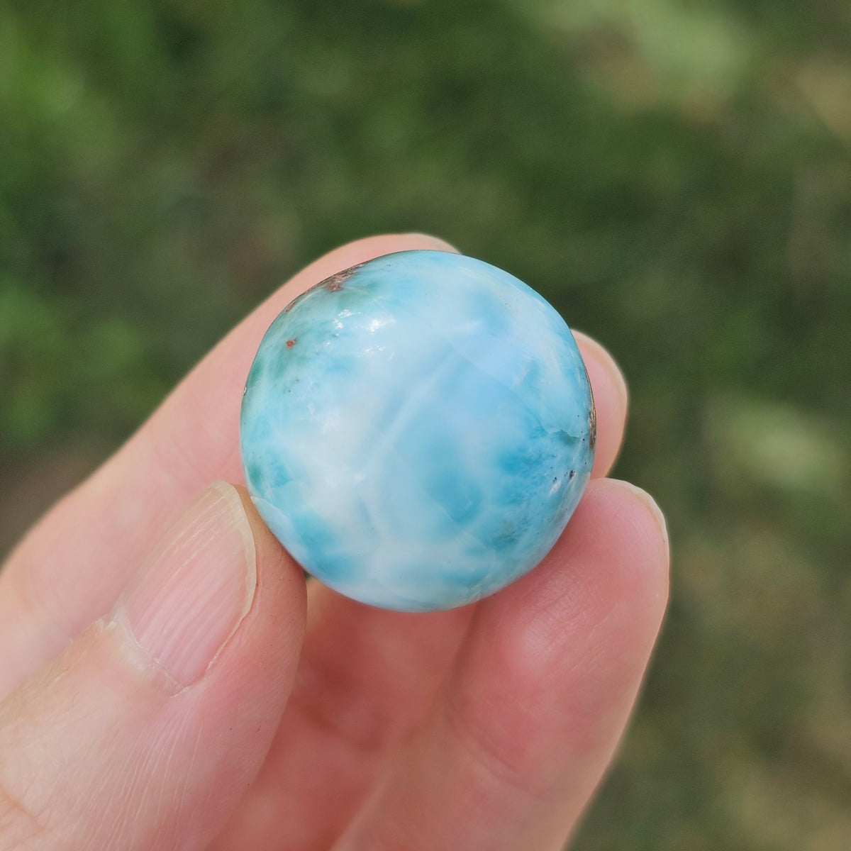 Grade AA natural blue Larimar sphere 16.8g 0.75 inch held in hand showing ocean wave patterns and natural hematite spots