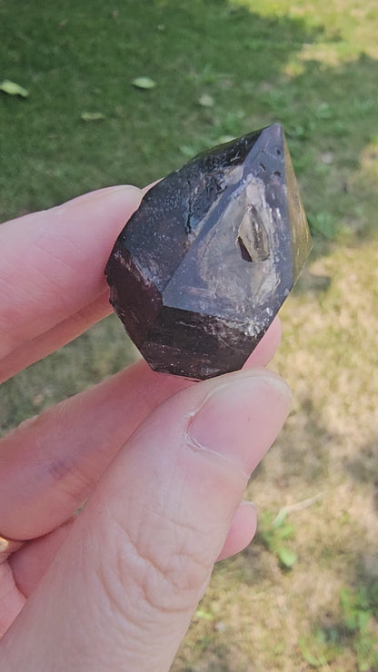 A close-up video of a hand rotating an 18g Brandberg amethyst crystal point in natural daylight, revealing its deep purple internal phantoms, clarity, and reflective facets.