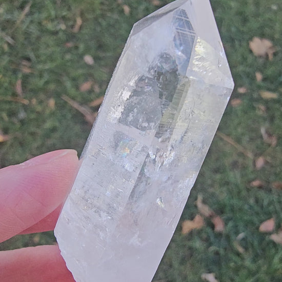 Close-up 360-degree video of a natural Colombian Lemurian Seed Quartz "Blade of Light" crystal. The footage highlights the specimen’s pristine optical clarity, distinct horizontal striations, and vivid internal rainbow flashes as it is rotated under natural light.